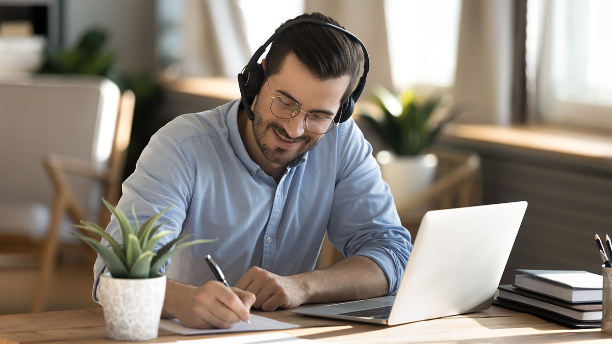 Smiling young Caucasian man in headphones glasses sit at desk work on laptop making notes.